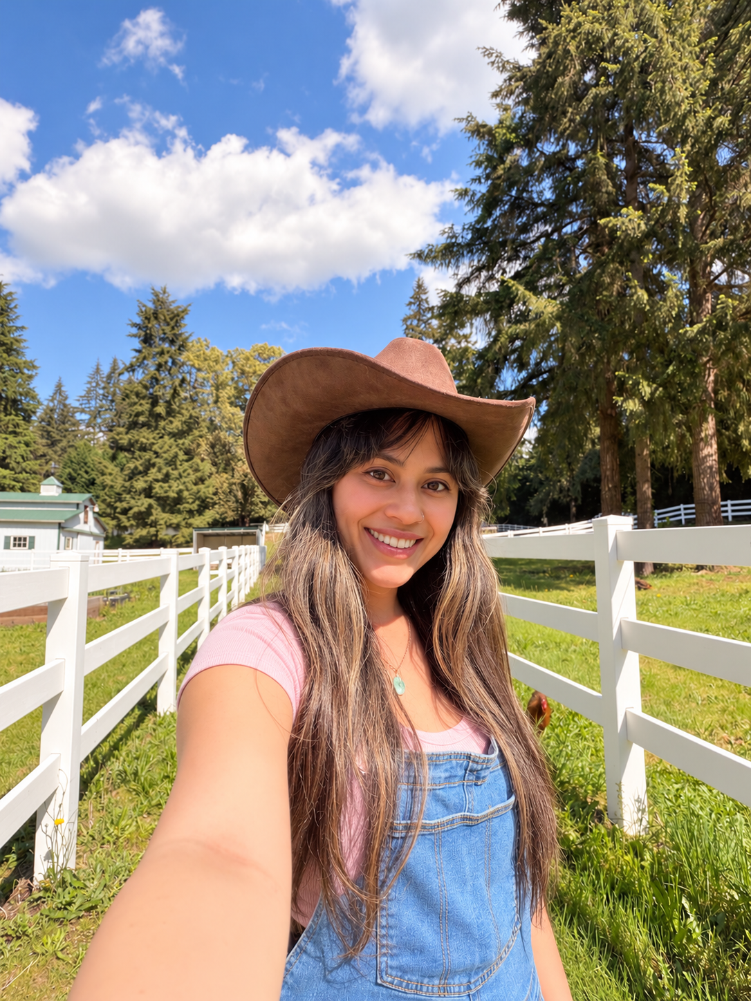 Kim smiling in a pink shirt, denim overalls, and cowboy hat beside a white farm fence